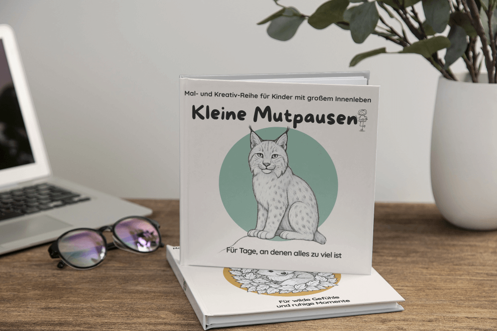kleine-mutpausen (19) Aufgestelltes Exemplar des Kinderbuchs „Kleine Mutpausen – Für Tage, an denen alles zu viel ist“ mit Luchs-Cover, dahinter ein weiteres Buch mit Fuchs-Cover, auf Tisch mit Brille und Pflanze.