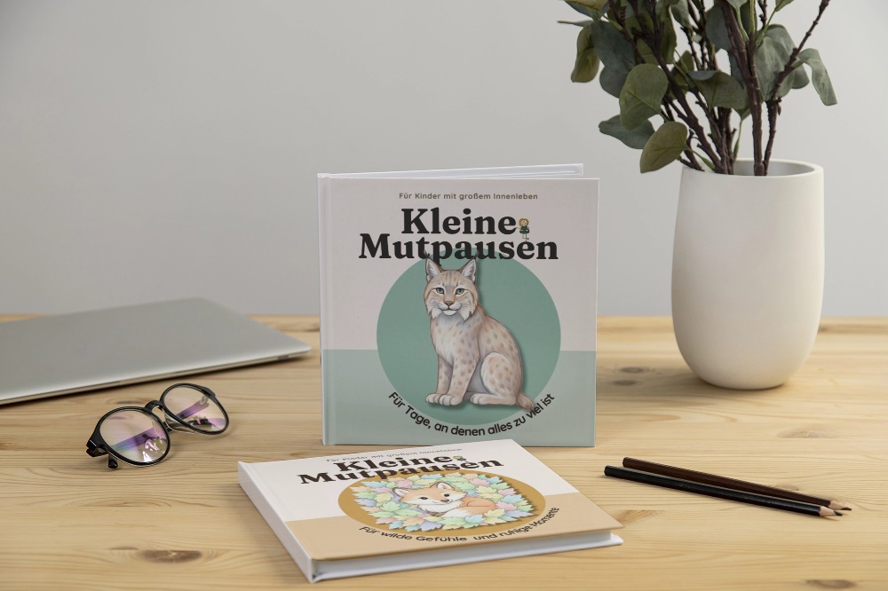 Aufgestelltes Exemplar des Kinderbuchs „Kleine Mutpausen – Für Tage, an denen alles zu viel ist“ mit Luchs-Cover, dahinter ein weiteres Buch mit Fuchs-Cover, auf Tisch mit Brille und Pflanze.